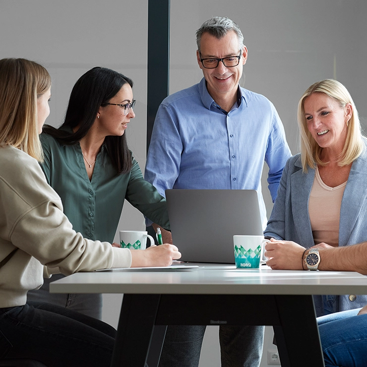 A group of smiling colleagues stand around a table and discuss things. 