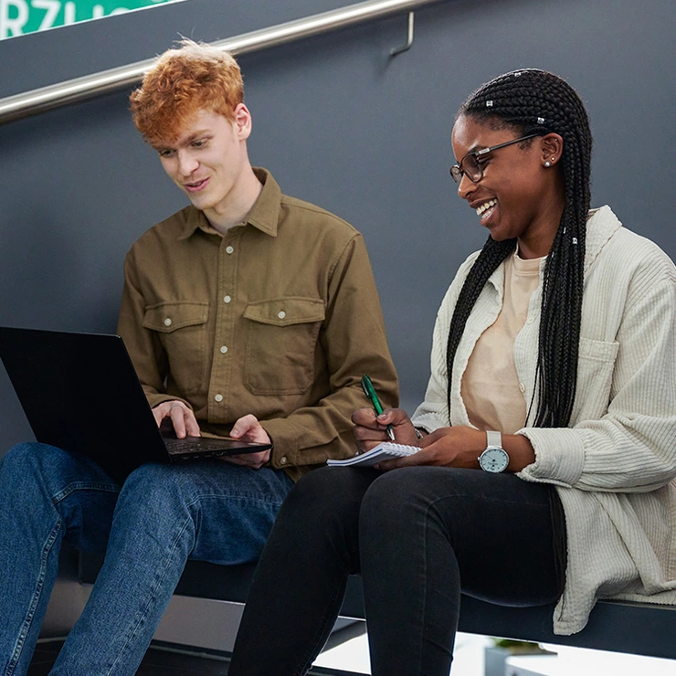 A young woman and a young man sit on a staircase with a laptop. 