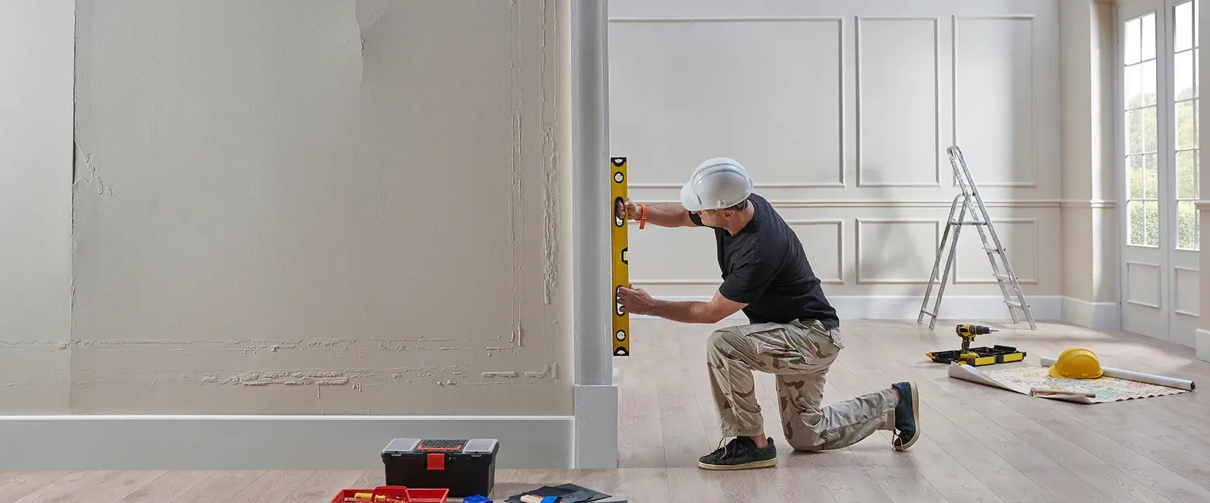 Construction worker in a flat holding a spirit level against the wall, with other tools lying around him.